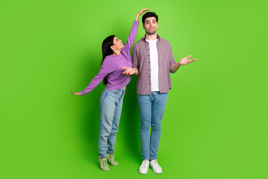 Portrait of a couple playfully comparing height on a green background, showcasing humor and connection in casual attire.