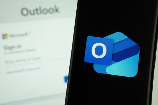 Dhaka, Bangladesh- 17 March 2025: microsoft outlook logo is displayed on a smartphone screen, with a stock graph webpage visible on a computer screen in the background.
