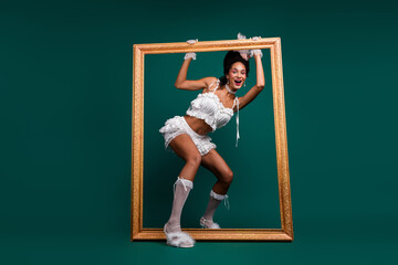 Young woman dressed in a playful baroque-style costume peers through a golden picture frame against...