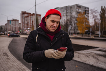 Fototapeta premium Man in a red beanie stands on a sidewalk looking at his smartphone in an urban setting during overcast weather