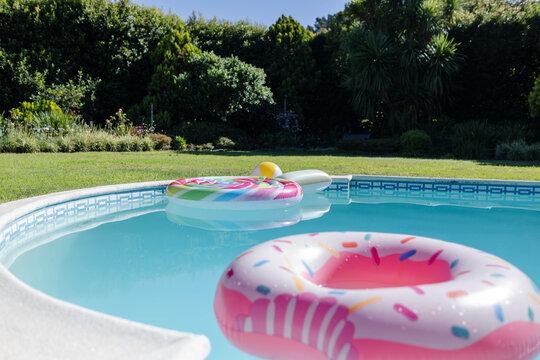 Naklejki Pink-and-white donut ring and lollipop swirl float drifting in clear pool water, framing green lawn