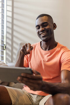 Smiling African American man interacting with tablet on couch in living room, with shuttered window