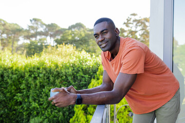 Leaning African American man holding coffee mug on balcony railing, wearing smartwatch, copy space