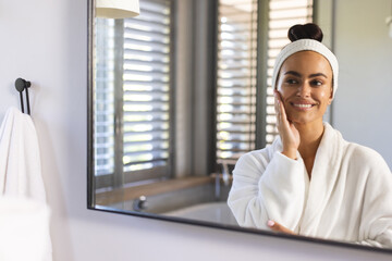 Smiling woman touching cheek in front of bathroom mirror, with bathrobe and headband, copy space
