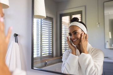 Applying cream woman in bathrobe pressing fingertips to cheeks in bathroom, with mirror, copy space