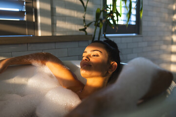 Reclining woman soaking in bathtub, with bubbles, plant and window blinds casting sunlight shadows