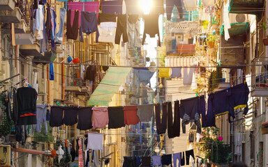 Street in Naples, Italy, with clothes hanging on lines between old buildings. The vibrant laundry contrasts with weathered walls, balconies, and sunlit facades, authentic southern Italian urban scene