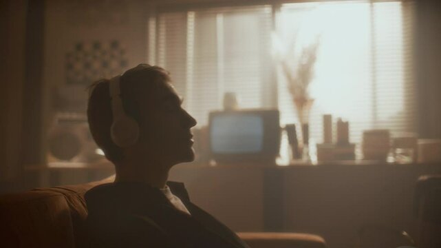 Young relaxed man listening to music with headphones, leaning back on couch and moving his head to rhythm in warmly lit room with retro-style decor. Side view