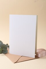 Blank greeting card, envelope, eucalyptus leaves and ribbon on beige background, closeup