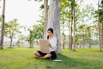 Bathed in warm golden light beneath a tree, a smiling female student immerses herself in her...