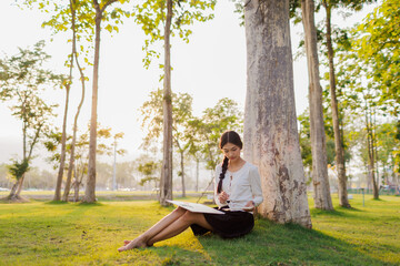 Bathed in warm golden light beneath a tree, a smiling female student immerses herself in her...