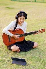 Fototapeta premium As golden rays of the setting sun touch the grass, an Asian woman sits quietly in a public park, playing a sweet melody on her guitar, letting the music blend with the evening breeze