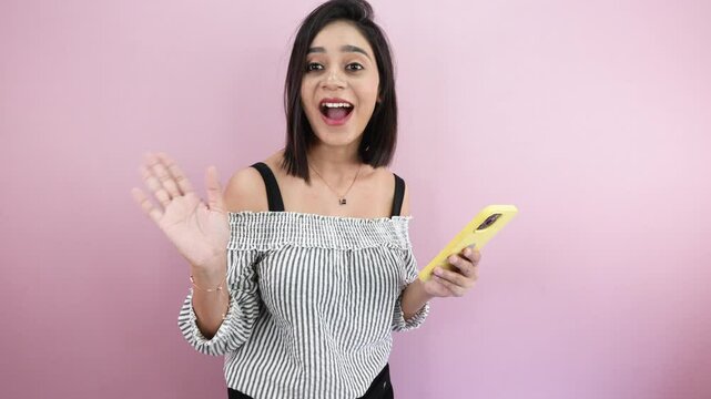 South Asian young woman holding smartphone, looking at camera and saying hi with waving hand gesture