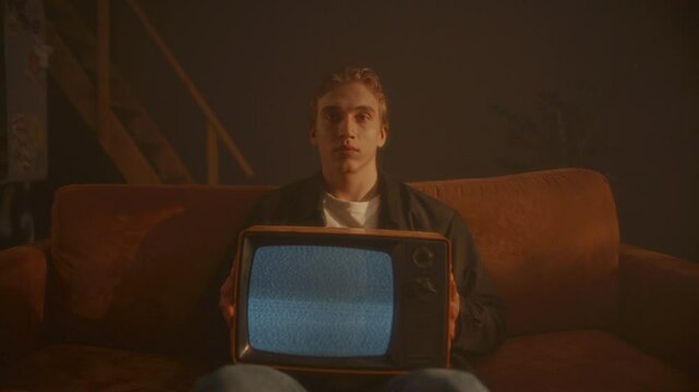 Young man sitting on sofa in dimly lit room, holding retro TV set with static screen and looking directly into camera with blank expression. Zoom-out, video portrait