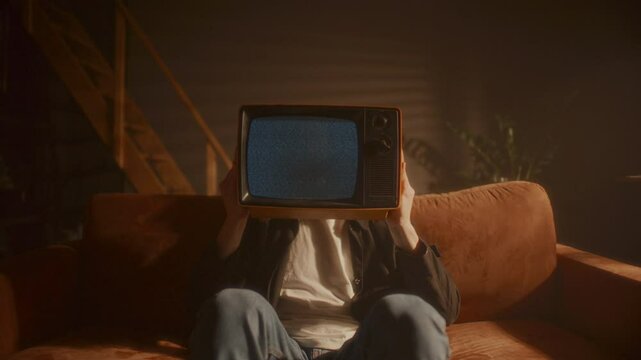 Young man sitting on sofa in warmly lit living room, holding vintage analog TV with static screen in front of his face. Zoom shot