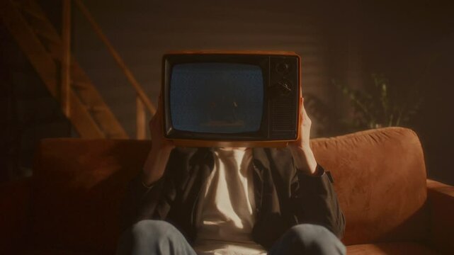 Man sitting on couch, holding retro TV set with static screen visible in front of his face in cozy living room with warm light. Zoom shot