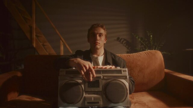 Young handsome man sitting on sofa in cozy warmly lit room, looking directly into camera and inserting cassette tape into retro boombox. Zoom-out, video portrait