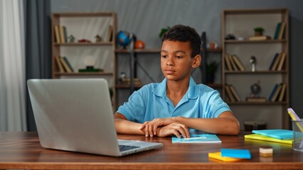 Young african american child boy studying at home, kid sits at desk attends school class online on laptop, looks at teacher on screen and listens information.
