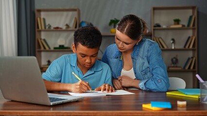Obraz premium Young african american child boy studying at home, kid and mother at desk, boy doing homework writing in notebook, mother helping and checking the process.