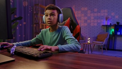 Young african american child boy spending time at home, kid streamer gamer in headphones sits at...