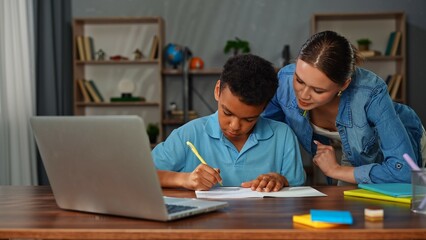 Young african american child boy studying at home, kid and mother at desk, boy doing homework writing in notebook, mother helping and checking the process.