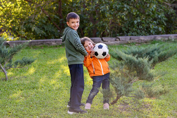 Happy siblings, boy and girl, playing with soccer ball in park on sunny day. Brother and toddler sister playing outdoors with football ball against green trees. Children outdoor activity concept