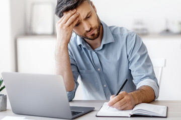 Serious Millennial Businessman Taking Notes And Working On Laptop Computer In Modern Office, Busy Entrepreneur Planning Business Schedule, Writing IN Notepad, Sitting At Desk On Workplace, Free Space
