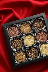 Delicious flower shaped chocolate bonbons in box on red fabric background, top view