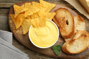 Tasty cheese dipping sauce in bowl, nacho chips and croutons on wooden table, top view