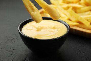 Dipping French fries into cheese sauce at black table, closeup