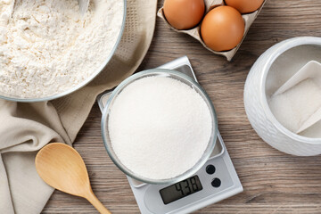 Digital kitchen scale with bowl of sugar among other products on wooden table, flat lay