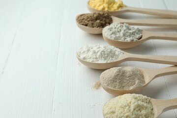 Different types of flour in spoons on white wooden table, closeup. Space for text
