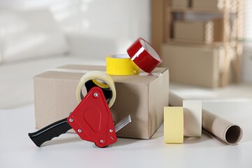 Dispenser with adhesive tape rolls and cardboard box on desk indoors