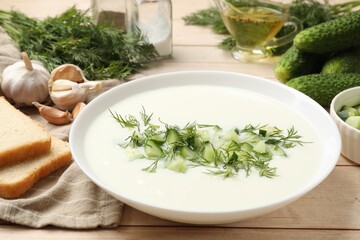 Tasty cucumber soup in bowl and ingredients on light wooden table, closeup