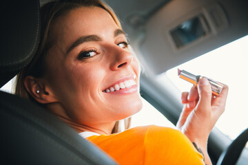 Young woman applying lipstick while sitting in a car on a sunny day, smiling confidently with a...