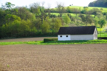 Grünes Landschaftspanorama mit braunem Ackerfeld, Stallgebäude von Bauernhof und Bergwiese mit Bäumen auf dem Land am Morgen im Frühling