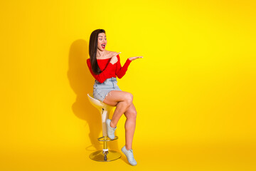 Happy young woman in a vibrant outfit pointing with enthusiasm against a bright yellow background, seated stylishly on a modern stool