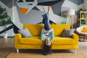 Charming woman relaxing upside down on a yellow sofa in a bright modern home during a peaceful day...