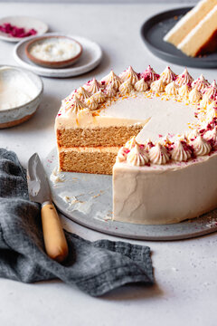 A sliced vanilla cake with cream frosting and decorative toppings on a plate, accompanied by a knife and linen napkin.