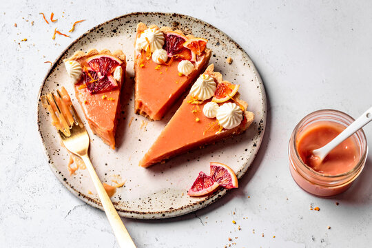 Three slices of blood orange pie with whipped cream and orange zest on a speckled plate, accompanied by a jar of blood orange curd.