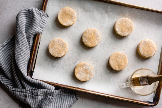 An overhead view of a baking tray with uncooked round cookies on parchment paper, alongside a brush and a cup of liquid for glazing.