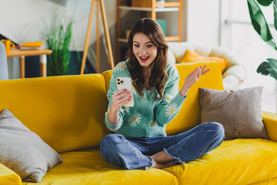 Charming woman sitting on a yellow sofa with a lively smile, holding a phone in her hands, enjoying her leisure time at home