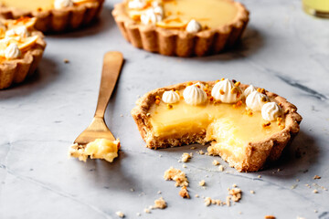 A lemon tart with a bite taken out, topped with cream and sprinkles, sits on a marble surface with a fork beside it.
