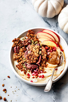 A bowl of yogurt topped with granola, sliced apples, pecans, pumpkin seeds, pomegranate seeds, and a dollop of nut butter, on a marble surface with small white pumpkins in the background.