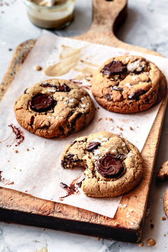 An assortment of freshly baked cookies with chocolate chunks on a wooden board, accompanied by a small jar of creamy spread.