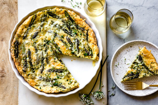 An asparagus and cheese quiche with a slice served on a plate, accompanied by glasses of white wine.