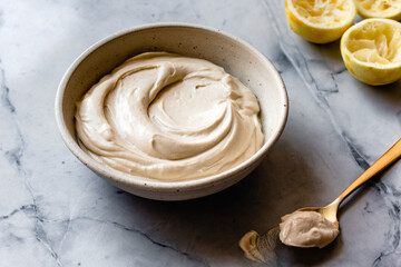 A bowl of creamy sauce with a spoon and squeezed lemons on a marble surface.