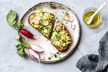 An overhead shot of avocado toast with radish slices on a speckled plate, accompanied by fresh radishes and a jar of olive oil.