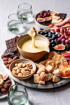An assortment of snacks including a bowl of melted cheese, crackers, bread, figs, grapes, almonds, and chocolate, with a person dipping bread into the cheese.