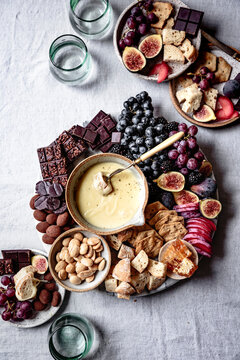 An elegant charcuterie board featuring a variety of cheeses, figs, grapes, almonds, chocolate, bread, and crackers, with a pot of creamy cheese dip on a textured white tablecloth.